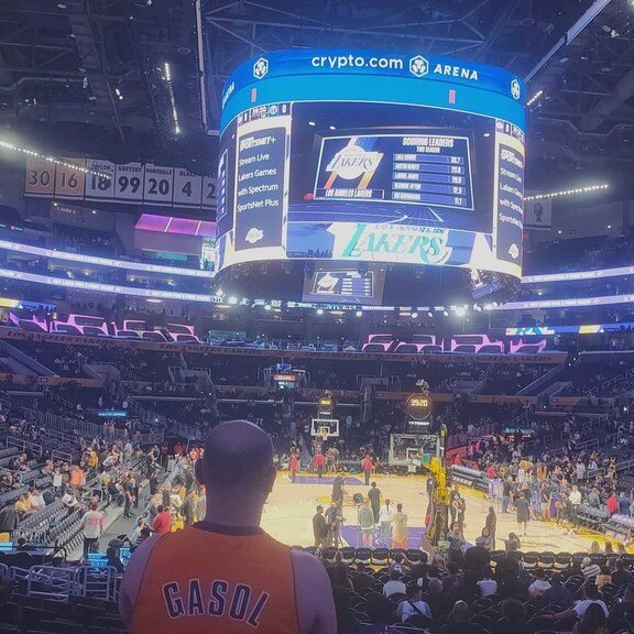 Espectador de espaldas con una camiseta de los Lakers de Pau Gasol viendo un partido de la NBA en un pabellón lleno con pantallas gigantes. 
